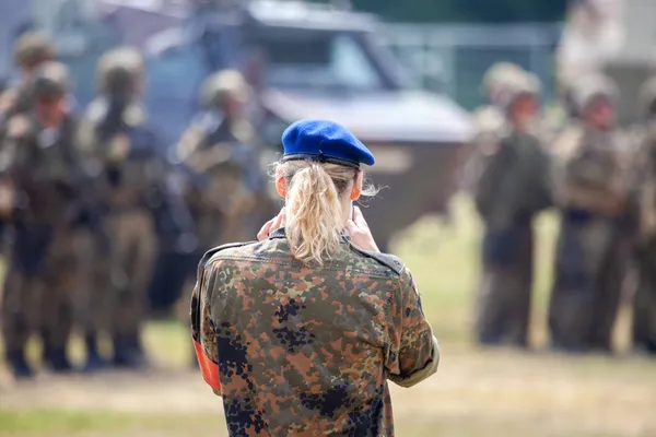 Eine Soldatin in Rückansicht, mit blonden Haaren, Pferdeschwanz, blauer Kappe und Tarnkleidung steht auf einem Übungsplatz. Im Hintergrund sind unscharf weitere Soldat:innen zu erkennen.